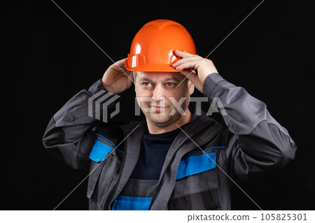 A young man construction worker in a safety helmet and work uniform on a black background A young man construction worker in a safety helmet and work uniform on a black background 105825301
