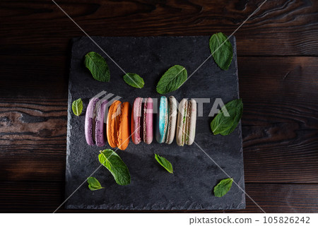 Macaroons of different colors and leaves lie on a slate bottom. Serving sweet macaroons. Sweet French dessert. Macaroons of different colors and leaves lie on a slate bottom. Serving sweet macaroons. Sweet French dessert. 105826242