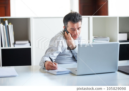 Consultant, businessman with smartphone on a call and with laptop writing notes in a notebook at his office. Customer service or online communication, networking and male person a cellphone at desk Consultant, businessman with smartphone on a call and with laptop writing notes in a notebook at his office. Customer service or online communication, networking and male person a cellphone at desk 105829098