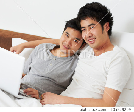 Portrait, laptop and bed with a gay couple watching a movie together while bonding in the morning. Love, happy or smile with an lgbt man and partner lying in their bedroom to relax while streaming 105829209
