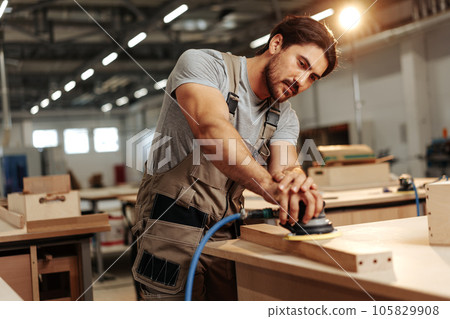 Young carpenter sanding wood piece in workshop in furniture factory 105829908