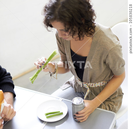 Health, diet and woman eating a celery stick for weightloss, wellness or vegetarian snack. Disappointment, upset and young female person with a green vegetable and beverage by a dining table at home. 105830218