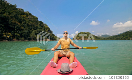 Man with sunglasses and hat rows pink plastic canoe along sea ag 105830363