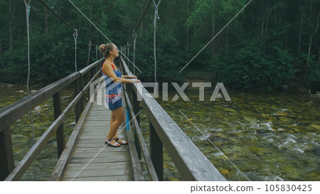 Woman Walking Along Suspension Bridge Alone in Picturesque Green Forest Setting. Woman Walking Along Suspension Bridge Alone in Picturesque Green Forest Setting. 105830425