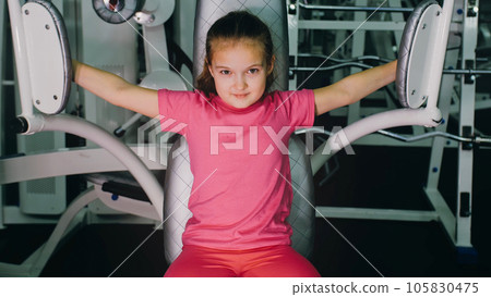 Fit Athletic Baby Girl Work out in Butterfly Pectoral Machine for Chest, Doing Her Fitness Exercise. Children Activity Training in Modern Gym. 105830475