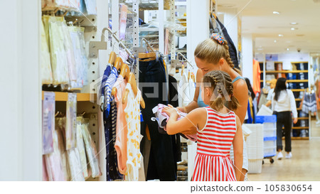 Young mother and child chooses clothes in sports clothing boutique. Cheerful child and mother choosing sports clothes in retail store. Mother and daughter choose looking for clothes in shopping mall. 105830654