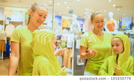 Young mother and child chooses clothes in sports clothing boutique. Cheerful child and mother choosing sports clothes in retail store. Mother and daughter choose looking for clothes in shopping mall. 105830779