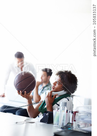 Hes ready to hit the courts. A young man playing with a basketball while sitting in the office. Hes ready to hit the courts. A young man playing with a basketball while sitting in the office. 105833137