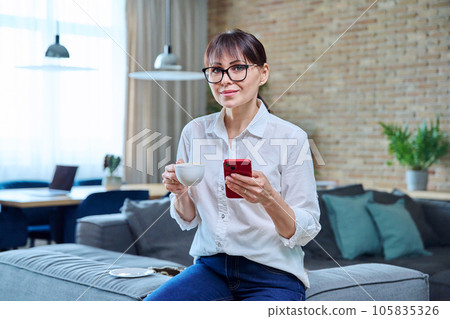 Middle aged woman using smartphone, having cup of coffee at home Middle aged woman using smartphone, having cup of coffee at home 105835326