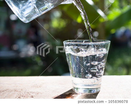Pouring Mineral Pure Water into Glass on Wooden Table with Over Light Day and Blur Green Nature Outdoor Background,Fresh Splash Ice Liquid Drink in Cristal Glass,Purified Food for Diet and Good Health 105836078