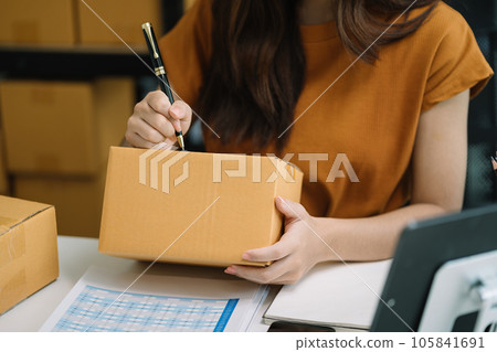 woman start up small business owner writing address on cardboard box at workplace.small business entrepreneur SME or freelance America woman working with box at home. woman start up small business owner writing address on cardboard box at workplace.small business entrepreneur SME or freelance America woman working with box at home. 105841691
