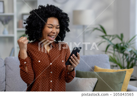 African american woman happy received online notification, good news in notification, woman sitting at home on couch in living room and celebrating victory success, holding hand up gesture of triumph 105843565
