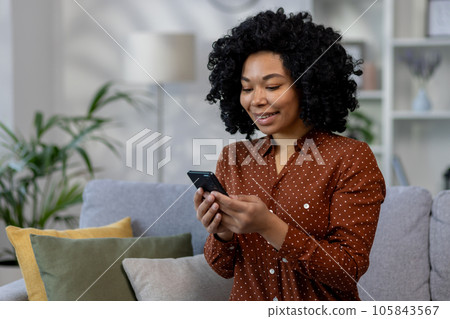 Young beautiful African American woman sitting on sofa at home, woman with curly hair holding phone in hands, using online application in living room browsing internet pages and typing messages Young beautiful African American woman sitting on sofa at home, woman with curly hair holding phone in hands, using online application in living room browsing internet pages and typing messages 105843567