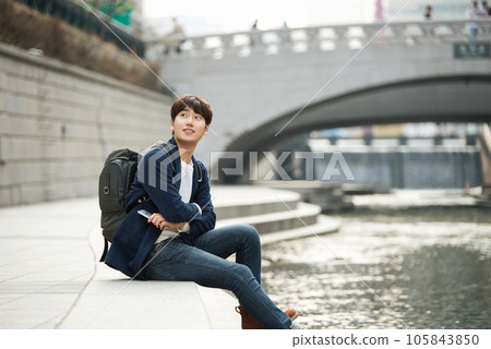 A young man relaxing by the river in the city and taking pictures with his smartphone 105843850