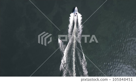 A young man rides a wakeboard. A sports guy rides on the water on a board behind a boat. Top view from a drone. 105848451