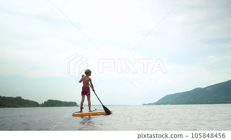 A boy on a sapsurf swims along the river against the background of the river. A sports child swims while standing on a sap. View from the back. A boy on a sapsurf swims along the river against the background of the river. A sports child swims while standing on a sap. View from the back. 105848456