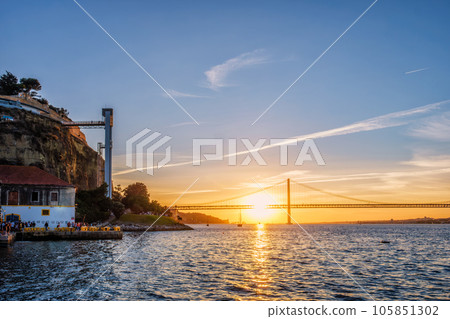 View of 25 de Abril Bridge famous tourist landmark of Lisbon over Tagus river and Boca do Vento Elevator and seaside restaurants with people at the sunset. Lisbon, Portugal 105851302