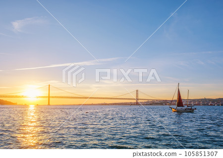 View of 25 de Abril Bridge famous tourist landmark of Lisbon connecting Lisboa and Almada over Tagus river with tourist yacht silhouette at sunset. Lisbon, Portugal 105851307