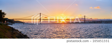 Panorama of 25 de Abril Bridge famous tourist landmark of Lisbon connecting Lisboa and Almada on Setubal Peninsula over Tagus river with tourist yacht silhouette at sunset. Lisbon, Portugal 105851331