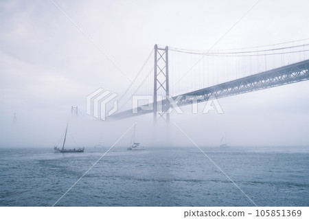 View of 25 de Abril Bridge famous tourist landmark of Lisbon connecting Lisboa and Almada in heavy fog mist wtih yacht boats passing under. Lisbon, Portugal 105851369