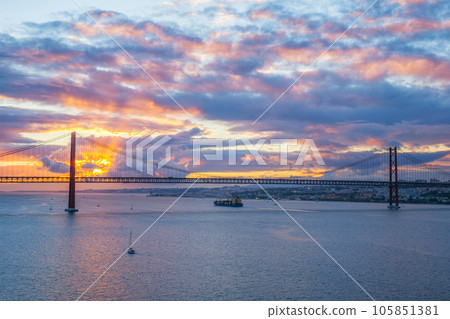 View of 25 de Abril Bridge famous tourist landmark of Lisbon over Tagus river with tourist yacht boats and cargo container ship on sunset. Lisbon, Portugal 105851381