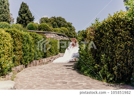 Brunette runs white dress park. A beautiful woman with long brown hair and a long white dress runs along the path along the beautiful bushes in the park, rear view 105851982