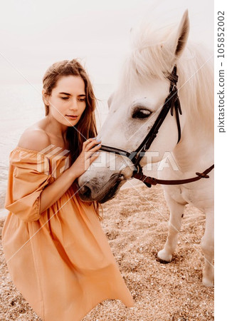 A white horse and a woman in a dress stand on a beach, with the sky and sea creating a picturesque backdrop for the scene. A white horse and a woman in a dress stand on a beach, with the sky and sea creating a picturesque backdrop for the scene. 105852002