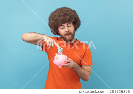 Portrait of positive happy man with Afro hairstyle wearing orange T-shirt investing, putting dollar banknote into piggy bank, money savings. Indoor studio shot isolated on blue background. Portrait of positive happy man with Afro hairstyle wearing orange T-shirt investing, putting dollar banknote into piggy bank, money savings. Indoor studio shot isolated on blue background. 105856896