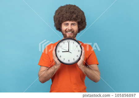 Portrait of satisfied delighted man with Afro hairstyle wearing orange T-shirt holding in hands big wall clock looking at camera with happy expression. Indoor studio shot isolated on blue background. Portrait of satisfied delighted man with Afro hairstyle wearing orange T-shirt holding in hands big wall clock looking at camera with happy expression. Indoor studio shot isolated on blue background. 105856897