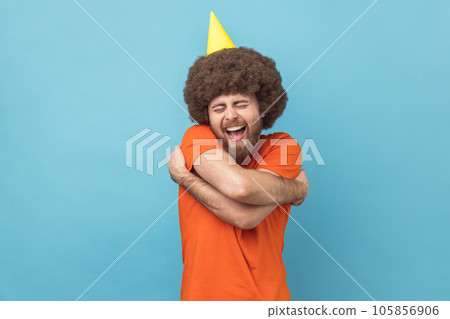 Man with Afro hairstyle in party cone embracing himself with expression of pleasure, happy to celebrate holiday alone, self-love concept. Indoor studio shot isolated on blue background. Man with Afro hairstyle in party cone embracing himself with expression of pleasure, happy to celebrate holiday alone, self-love concept. Indoor studio shot isolated on blue background. 105856906