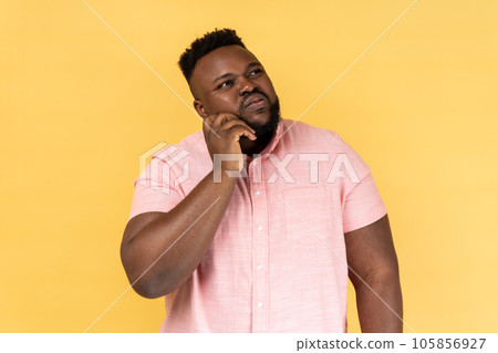 Portrait of pensive young adult bearded man wearing pink shirt touching chin and thinking about important question, making hard decision. Indoor studio shot isolated on yellow background. Portrait of pensive young adult bearded man wearing pink shirt touching chin and thinking about important question, making hard decision. Indoor studio shot isolated on yellow background. 105856927