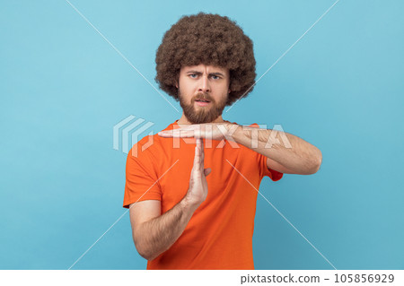 Portrait of serious man with Afro hairstyle wearing orange T-shirt showing timeout gesture, tired of lot of homework, extra classes. Indoor studio shot isolated on blue background. Portrait of serious man with Afro hairstyle wearing orange T-shirt showing timeout gesture, tired of lot of homework, extra classes. Indoor studio shot isolated on blue background. 105856929