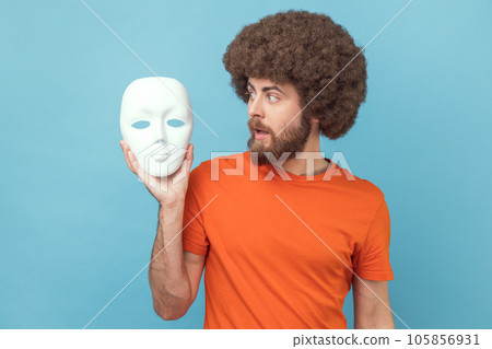 Portrait of man with Afro hairstyle wearing orange T-shirt looking at white mask in hands with attentive look, trying to understand hiding personality. Indoor studio shot isolated on blue background. 105856931
