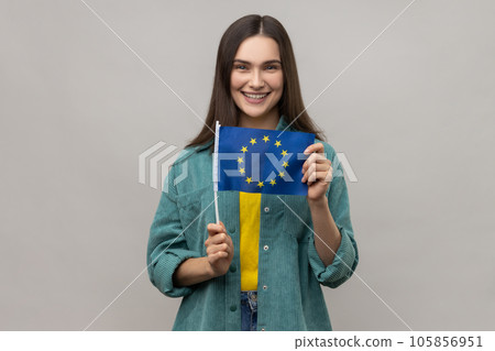 EU association. Happy tourist woman holding flag of European Union, immigration and traveling in Europe, wearing casual style jacket. Indoor studio shot isolated on gray background. EU association. Happy tourist woman holding flag of European Union, immigration and traveling in Europe, wearing casual style jacket. Indoor studio shot isolated on gray background. 105856951
