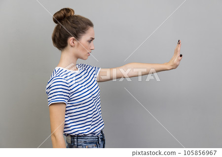Side view portrait of woman wearing striped T-shirt showing stop gesture, warning with raised palm, showing ban, block sign with hand. Indoor studio shot isolated on gray background. 105856967