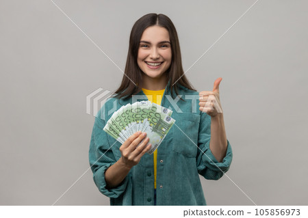 Smiling dark haired woman holding many euro banknotes and showing thumb up to camera, satisfied of winning lottery, wearing casual style jacket. Indoor studio shot isolated on gray background. Smiling dark haired woman holding many euro banknotes and showing thumb up to camera, satisfied of winning lottery, wearing casual style jacket. Indoor studio shot isolated on gray background. 105856973