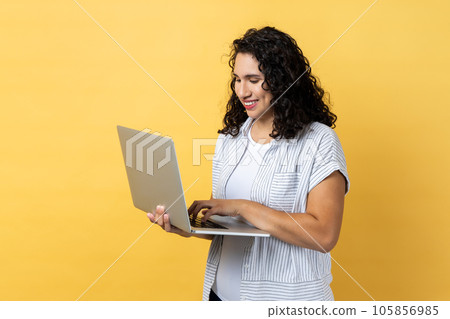 Portrait of cute positive optimistic woman with dark wavy hair working on laptop, looking at display screen with pleasant expression. Indoor studio shot isolated on yellow background. Portrait of cute positive optimistic woman with dark wavy hair working on laptop, looking at display screen with pleasant expression. Indoor studio shot isolated on yellow background. 105856985