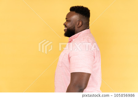 Side view of happy satisfied young adult man wearing pink shirt standing with positive facial expression, looking ahead with toothy smile. Indoor studio shot isolated on yellow background. Side view of happy satisfied young adult man wearing pink shirt standing with positive facial expression, looking ahead with toothy smile. Indoor studio shot isolated on yellow background. 105856986