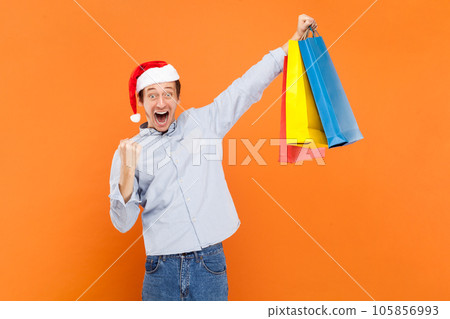 Portrait of extremely happy young adult man clenched fist and holding shopping bags, celebrating successful shopping, wearing light blue shirt and Santa Claus hat. Indoor studio shot isolated on 105856993