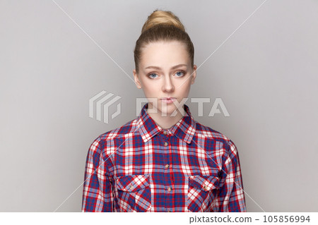 Portrait of sad upset depressed woman with bun hairstyle standing looking at camera, being offended and stressed, wearing checkered shirt. Indoor studio shot isolated on gray background. 105856994