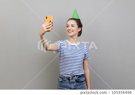 Portrait of positive woman blogger wearing striped T-shirt and party cone taking selfie, talking with followers, having video call. Indoor studio shot isolated on gray background. Portrait of positive woman blogger wearing striped T-shirt and party cone taking selfie, talking with followers, having video call. Indoor studio shot isolated on gray background. 105856996