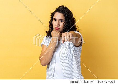 Portrait of aggressive woman with dark wavy hair holding clenched fists up, fighting with abuser, defence from domestic violence. Indoor studio shot isolated on yellow background. 105857001