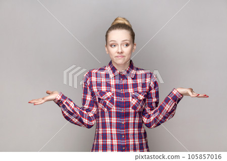 Portrait of uncertain confused attractive woman with bun hairstyle shrugging shoulders, dosen't know answer, wearing checkered shirt. Indoor studio shot isolated on gray background. Portrait of uncertain confused attractive woman with bun hairstyle shrugging shoulders, dosen't know answer, wearing checkered shirt. Indoor studio shot isolated on gray background. 105857016