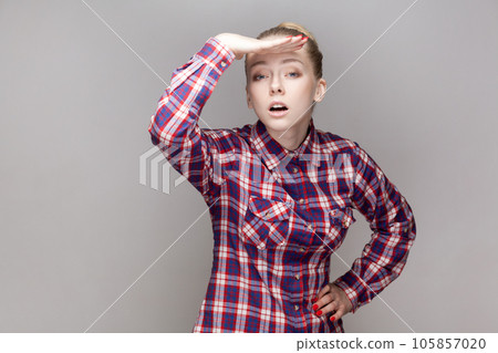 Portrait of attentive concentrated attractive woman with bun hairstyle looking far in distance with hand near forehead, wearing checkered shirt. Indoor studio shot isolated on gray background. Portrait of attentive concentrated attractive woman with bun hairstyle looking far in distance with hand near forehead, wearing checkered shirt. Indoor studio shot isolated on gray background. 105857020