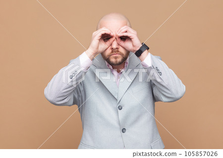 Portrait of serious concentrated bald bearded man standing raised arms and looking with binoculars gesture, wearing gray jacket. Indoor studio shot isolated on brown background. 105857026