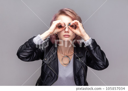 Portrait of attentive concentrated woman with short hairstyle standing making binocular gesture, looking far, wearing black leather jacket. Indoor studio shot isolated on grey background. Portrait of attentive concentrated woman with short hairstyle standing making binocular gesture, looking far, wearing black leather jacket. Indoor studio shot isolated on grey background. 105857034
