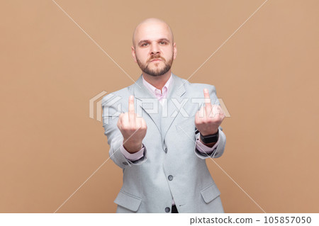 Portrait of crazy angry bald bearded man showing middle finger, rude gesture, face full of hatred and resentment, wearing gray jacket. Indoor studio shot isolated on brown background. 105857050