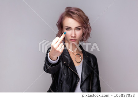 Portrait of serious rude woman with short hairstyle standing showing middle finger, impolite gesture, looking at camera, wearing black leather jacket. Indoor studio shot isolated on grey background. 105857058