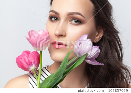Portrait of charming romantic brunette woman looking at camera, holding spring tulips, enjoying pink flowers, wearing striped dress. Indoor studio shot isolated on gray background. 105857059