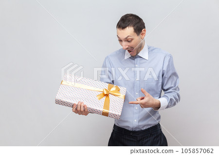 Portrait of surprised positive cheerful man holding wrapped present box, pointing at gift with amazed expression, wearing light blue shirt. Indoor studio shot isolated on gray background. 105857062